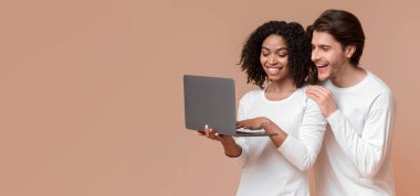 Çevrimiçi satış kavramı. Mutlu ırklar arası eşler dizüstü bilgisayarlarını tutuyor ve heyecanlı bir şekilde bilgisayar ekranına bakıyor, internet alışverişinden keyif alıyorlar.