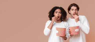 Korku filmi. Kovalardan patlamış mısır yiyen ve korkmuş yüz ifadesiyle kameraya bakan çok ırklı bir çift. Sarı arka plan.