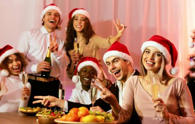 Noel Baba Şapkalı Neşeli Dostlar ellerinde şampanya kadehleri, evinde yeni yılı kutlayan kameraya gülümseyen zafer işareti.