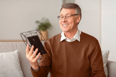 Neşeli, yaşlı bir adam elinde fotoğraf çerçevesiyle oturma odasında oturan aile portresine bakıyor. Seçici Odaklanma