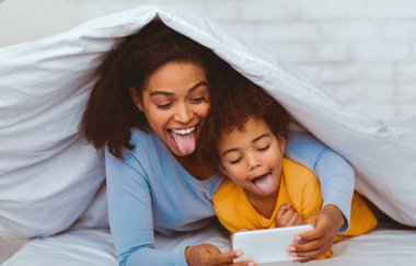 Neşeli anne ve kızı evde battaniyeyle yatakta uzanmış cep telefonu oyunları oynuyorlardı. Aile eğlencesi ve eğlence. Seçici Odaklanma