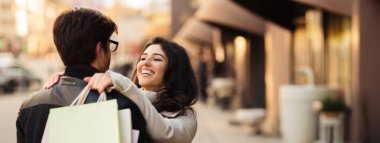 Bir çift gülümsüyor ve birbirlerine sarılıyor. Bahar zamanı kalabalık bir şehir caddesinde alışveriş torbaları tutarken. Kopya alanı.