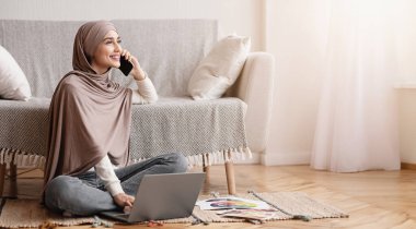Müslüman Kadınlar İçin Çalışma Fırsatları. tesettürlü gülümseyen Arap kız cep telefonuyla konuşuyor ve evde otururken dizüstü bilgisayar kullanıyor..