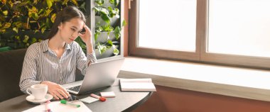 Genç bir kadın, masa başında otururken dizüstü bilgisayarına odaklanır. Etrafı bitkilerle ve doğal ışıklarla çevrilidir. Uzayı kopyalayabilir.