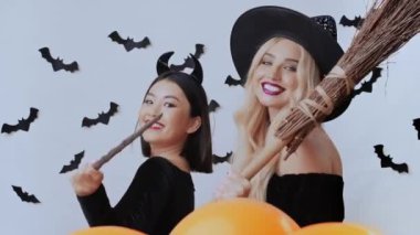 Cadılar Bayramı büyücüleri. İki çok ırklı kadın kostümlü, kameraya gülümseyen ve flört eden, sarışın süpürge tutan, Asyalı sihirli değnekle oynayan, stüdyo geçmişi olan