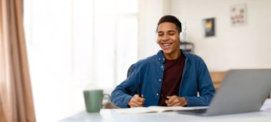 Genç siyah bir öğrenci, kulaklık takıyor, gülümsüyor ve parlak, rahat odasından online bir derse katılıyor. Dizüstü bilgisayarı ve not defteriyle bir masada oturuyor ve öğrenmeye odaklanıyor..