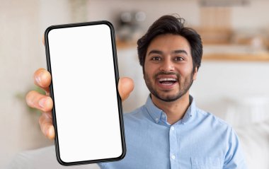 Hintli bir adam elinde boş bir akıllı telefon tutarken, heyecanını göstererek geniş bir gülümseme sergiliyor. Parlak ve davetkar dekorlarla çevrili şık bir evde duruyor..
