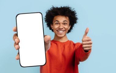 Kıvırcık saçlı Afro-Amerikalı genç adam boş bir akıllı telefonu tutarken gülümsüyor, basit, açık mavi bir arka plana karşı heyecan ve coşku gösteriyor..