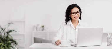Assistant. Portrait of happy mexican woman in glasses working on laptop, drinking coffee, copyspace
