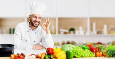 Profesyonel Aşçı. Gülümseyen aşçı, tamam işareti veriyor. Restorandaki mutfak masasının yanında duruyor.