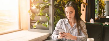 Genç bir kadın bir kafede oturmuş, etrafı yemyeşil bitkiler ve yumuşak güneş ışığıyla çevriliyken, akıllı telefonuna odaklanmış, boşluğu kopyalıyor.