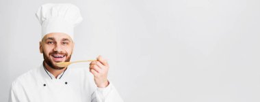 Chef Guy Tasting Dish Holding Spoon Smiling To Camera Posing Standing On Gray Studio Background. Gourmet Concept. Panorama, Empty Space