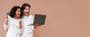 Joyful interracial couple rejoicing success with credit card and laptop, looking at computer screen and yelling with joy on yellow background