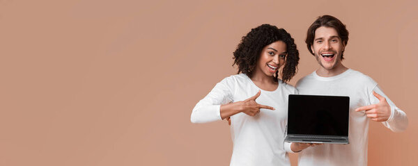 Cool website. Smiling multiracial couple holding laptop with black screen and pointing at it, posing together over yellow background with empty space