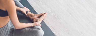 Bir kadın yoga minderinde bacak bacak üstüne atarak oturur, bacaklarını nazikçe uzatır ve sakin kapalı bir ortamda rahatlamaya odaklanır, kırpılmış, kopyalanmış bir yerde.