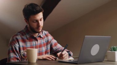Çevrimiçi eğitim, uzaktan öğrenme. Genç bir erkek öğrenci dizüstü bilgisayarda Webinar izleyip not alıyor, evde kahve masasında oturuyor.
