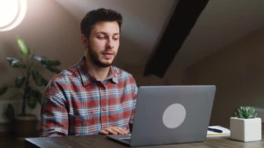 Uzaktan görüşme. Genç adam laptopta görüntülü sohbet yoluyla ortaklarla konuşuyor, ev ofisinde oturuyor, yavaş çekim.