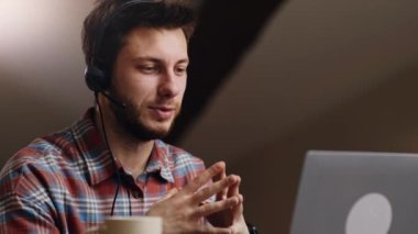 Çevrimiçi video konferansında kulaklık takan kendine güvenen adam, projeyi uzaktan işyerindeki jestlerle açıklıyor, evde dizüstü bilgisayarla çalışıyor.