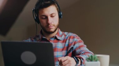 Yeni normal ve uzak öğrenim. Kulaklıklı genç adam internetten web sitesi izliyor ve notlar alıyor, evde uzaktan çalışıyor.