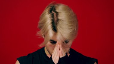 Bir insan elleri birbirine yaslanmış, başını eğip düşüncelere dalmış olarak ayakta durur. Çarpıcı kırmızı arkaplan, düşünceli ifadelerini ve sarı saçlarını vurguluyor ve canlı bir kontrast oluşturuyor..