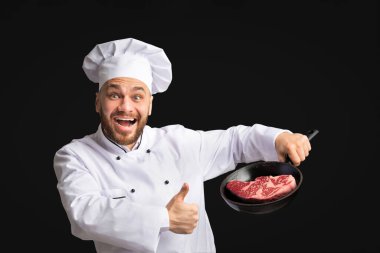 Yemek pişirmek. Mutlu Aşçı Tavayı Taze Et Bifteğiyle Tutuyor Ve Siyah Stüdyo Arkaplanında Ayakta Durarak İşaret Ediyor