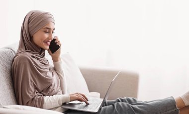 Eğlence konsepti için modern teknolojiler. tesettürlü gülümseyen Arap kız dizüstü bilgisayar kullanıyor ve cep telefonuyla konuşuyor, evdeki kanepede dinleniyor, boş yer var.