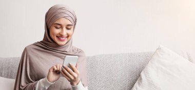 Mutlu Arap kadın tesettür içinde sosyal ağları kaydırıyor ya da evdeki akıllı telefondan fotoğraf izliyor, rahat bir koltukta oturuyor, boş bir alanda.