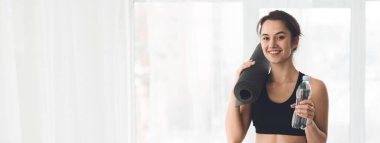 Genç bir kadın elinde yoga minderi ve su şişesi tutarken gülümsüyor, büyük pencereli parlak bir odada duruyor, boşluğu kopyalıyor.
