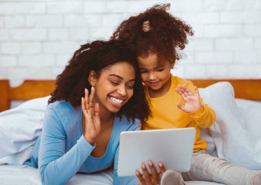 Black Mother And Daughter Using Digital Tablet Making Video Call And Waving Hello Lying In Bed At Home. Selective Focus