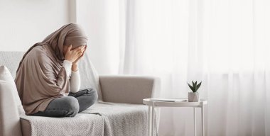 Depresyon kavramı. Üzgün, tesettürlü Müslüman bir kadın evdeki kanepede ağlıyor, yüzünü ellerde saklıyor, kendini çaresiz ve yalnız hissediyor, boş bir alan.
