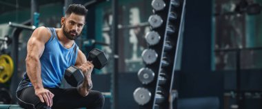 İyi donanımlı bir spor salonundaki bankta oturan bir adam, antremanını yaparken azimle bir dambılı kaldırmaya odaklanıyor, kopya alanlı panorama.