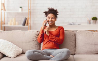 Telefon konuşması. Hafta sonları evde koltukta oturan cep telefonuyla konuşan gülümseyen Afro Kadın.