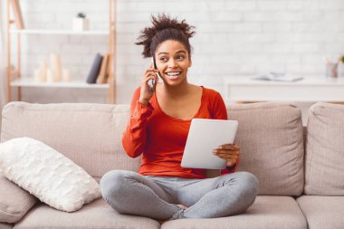 Gadget Lifestyle. Cheerful Black Woman Talking On Phone Using Digital Tablet Sitting On Sofa At Home.