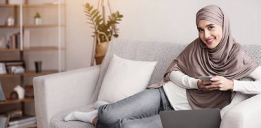 Tesettürlü neşeli Müslüman kız akıllı telefonuyla evde vakit geçiriyor ve kameraya bakıyor, tembelce hafta sonunun tadını çıkarıyor..