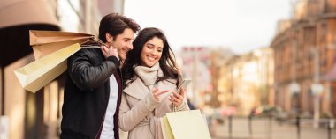 Neşeli çiftler cep telefonlarındaki sezonluk satışları kontrol ediyorlar, caddelerde birlikte yürüyorlar, alışverişleri tutuyorlar, kopyalama alanlarını tutuyorlar.