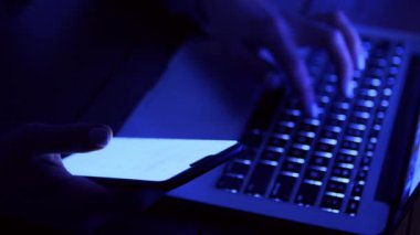Bir insan bir elinde akıllı telefon, diğer elinde ise soluk bir mavi ışıkla dizüstü bilgisayarda yazar..