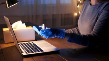 Mavi eldiven giyen biri ellerini dizüstü bilgisayar, mendil kutusu ve küçük bir şişeyle temizliyor..