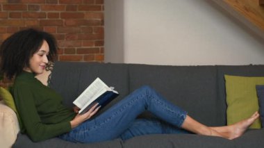 Keyifli boş zaman. Genç siyahi kadın kitap okuyor, evdeki kanepede dinleniyor, yan bakıyor.