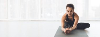 Bir kadın, güneşli bir stüdyoda minderin üzerinde oturup, esnekliğine ve dengesine odaklanarak yoga esneme hareketleri yapıyor.