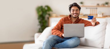 Genç bir Hintli adam bir kanepede rahatça oturuyor, elinde kredi kartı tutarken dizüstü bilgisayarını kullanırken gülümsüyor. Konforlu yaşam alanı rahat bir atmosfere sahip, online alışveriş için mükemmel..