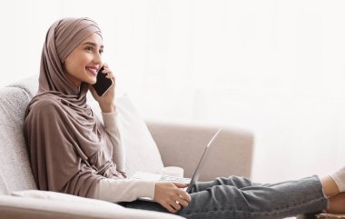 Smiling Muslim Girl In Hijab Having Phone Conversation And Using Laptop While Sitting On Sofa Against Window At Home, Free Space