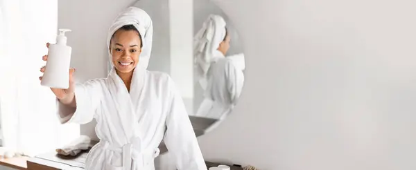 stock image African American woman in a white spa robe and towel happily presents a bottle of skincare product while surrounded by a clean, minimalistic bathroom with a mirror and natural light.