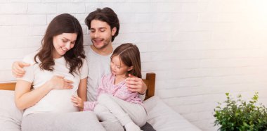 İkinci çocuk için hazır. Hamile kadın, kocası ve küçük kızı evde yatakta birbirlerine bağlanıyorlar, tatlı çocuk annelerinin karnına şefkatle dokunuyor.