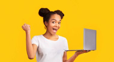 Başarımızı kutluyoruz. Stüdyoda dizüstü bilgisayarı tutan heyecanlı Afrikalı Amerikalı kızın portresi. Panorama