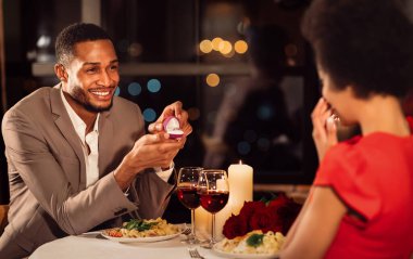 Marry me. Loving afro man proposing to girlfriend holding engagement wedding ring dating in fancy restaurant. Selective focus clipart