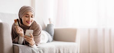 İnternetten alışveriş konsepti. Akıllı telefon ve kredi kartıyla tesettürlü neşeli Arap kadın evdeki koltukta dinleniyor, internetten mal alıyor, boş yer arıyor.