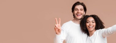 Happy Interracial Couple Taking Selfie. Cheerful Guy Showing Peace Sign While His Afro Girlfriend Capturing Photo, Yellow Background With Free Space
