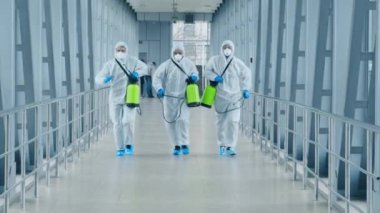 Koruyucu giysiler ve maskeli üç kişi, sağlık krizi sırasında binayı sterilize etmek için dezenfektan spreyleri taşıyan gösterişli bir koridorda koşuşturuyorlar..