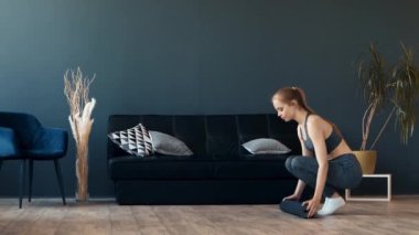 Genç bir kadın şık bir oturma odasındaki yoga minderinde çömelme gösterisi yapıyor. Mekanda minimalist kanepe ve dekoratif bitkiler de dahil olmak üzere modern mobilyalar yer alıyor..