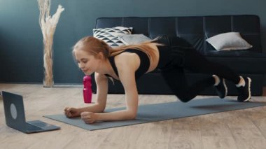 Genç bir kadın evinde yoga minderinde tahta egzersizi yapıyor. Dizüstü bilgisayarındaki antrenmanı yakından takip ediyor. Konforlu bir oturma odasına odaklanmış durumda..